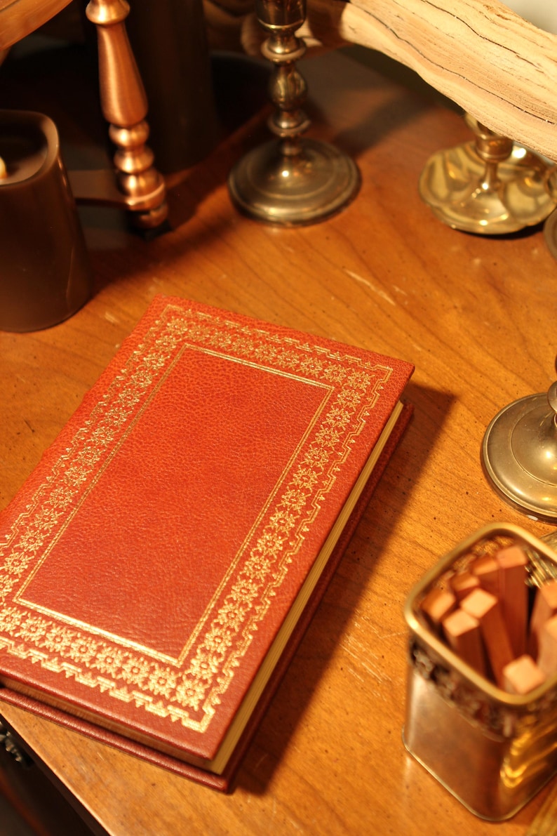 May include: A red leather-bound book with gold decorative trim. The book is on a wooden surface, alongside a candle, brass candlesticks, and a small container of matches. The book's cover has a textured appearance.