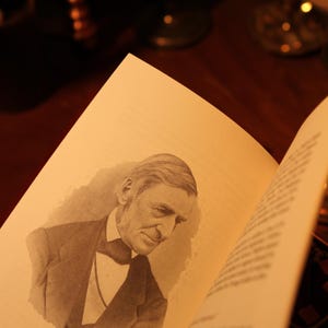 May include: An open book with a sepia-toned illustration of Ralph Waldo Emerson, with the text "Ralph Waldo Emerson (1803-1882)". The book is on a dark wooden surface, with blurred objects in the background.