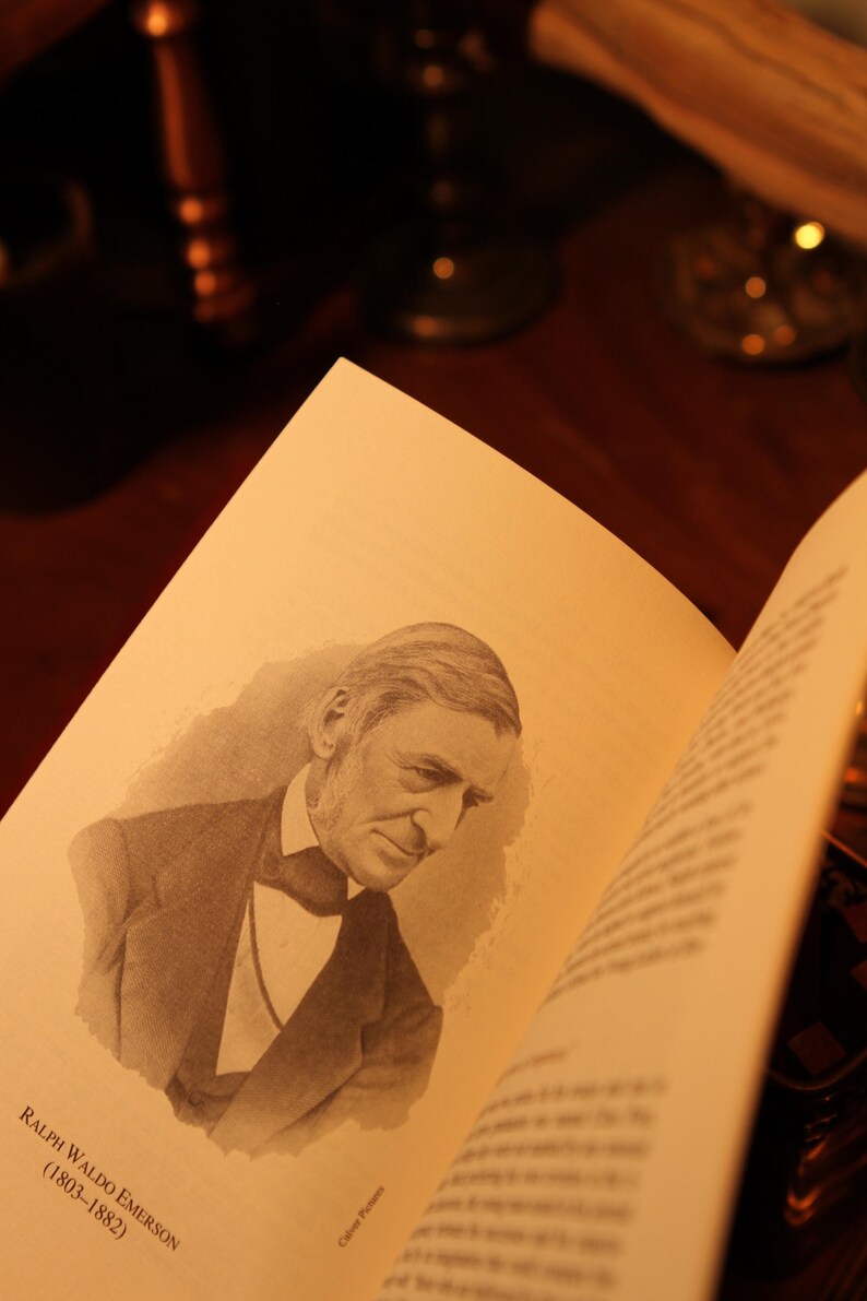 May include: An open book with a sepia-toned illustration of Ralph Waldo Emerson, with the text "Ralph Waldo Emerson (1803-1882)". The book is on a dark wooden surface, with blurred objects in the background.