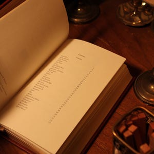 May include: An open book with a red cover, displaying a list of poems on a cream-colored page. The book is on a wooden surface, with a small metal container and candle holders in the background.