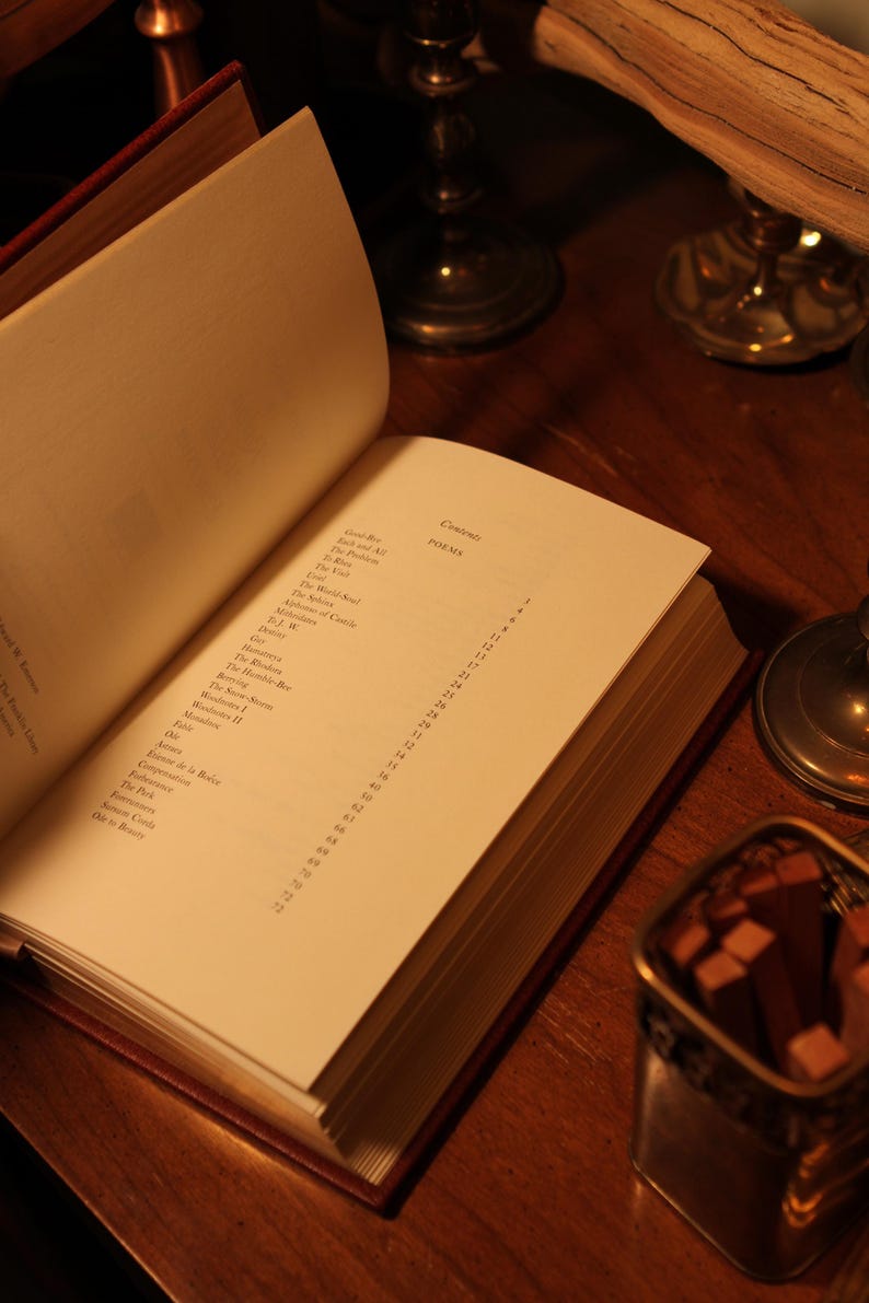 May include: An open book with a red cover, displaying a list of poems on a cream-colored page. The book is on a wooden surface, with a small metal container and candle holders in the background.