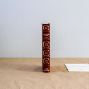 May include: A hardcover book with a burgundy leather cover and gold lettering. The spine features decorative gold accents and the title "Ruth Wader Ginsburg" and "Found". A small white card is next to the book.