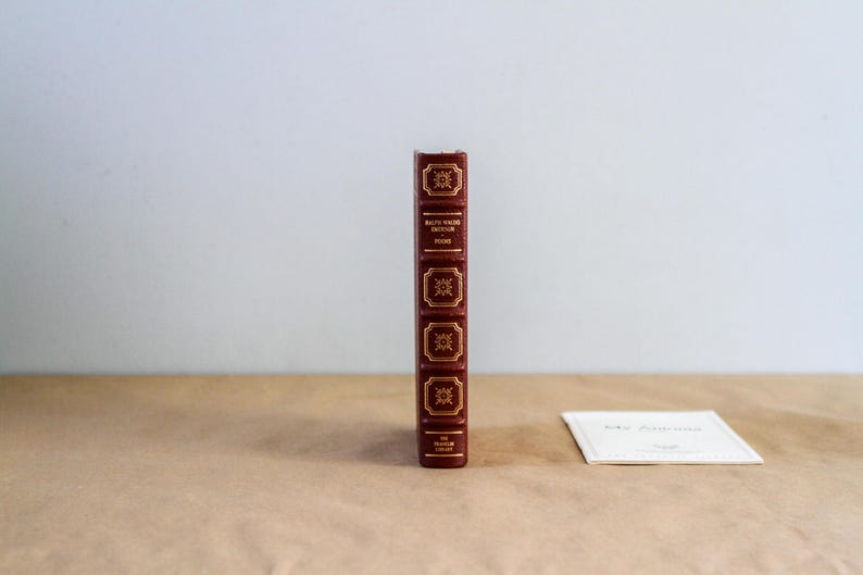May include: A hardcover book with a burgundy leather cover and gold lettering. The spine features decorative gold accents and the title "Ruth Wader Ginsburg" and "Found". A small white card is next to the book.