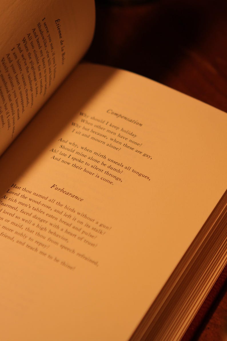 May include: Open book with pages displaying printed text. The text includes the title "Compensation" and the poem "Forbearance". The pages are a warm, off-white color, and the book is open on a wooden surface.