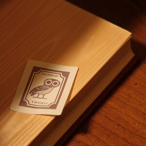 May include: A close-up of a book with a light brown cover and a decorative card. The card features an owl illustration and the text "TALISMAN" and "BOOKS" in a dark purple color. The book is on a wooden surface.