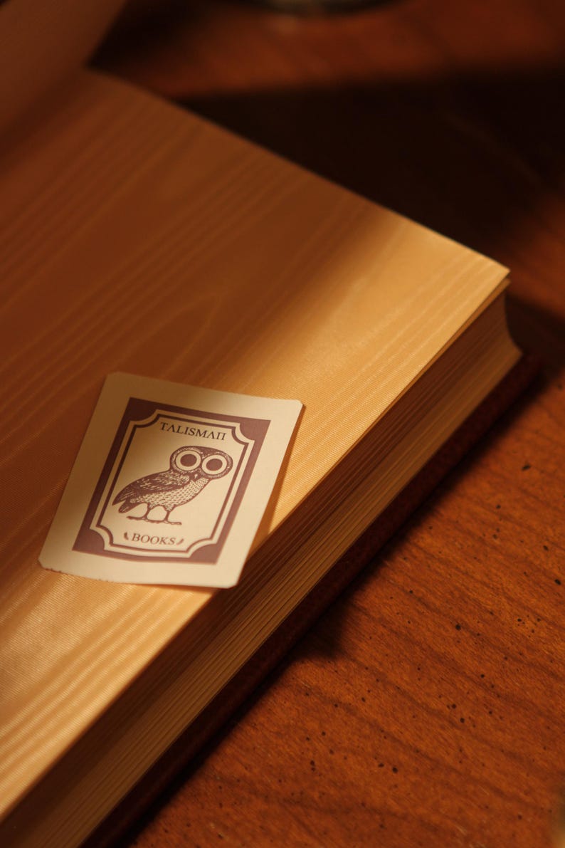 May include: A close-up of a book with a light brown cover and a decorative card. The card features an owl illustration and the text "TALISMAN" and "BOOKS" in a dark purple color. The book is on a wooden surface.