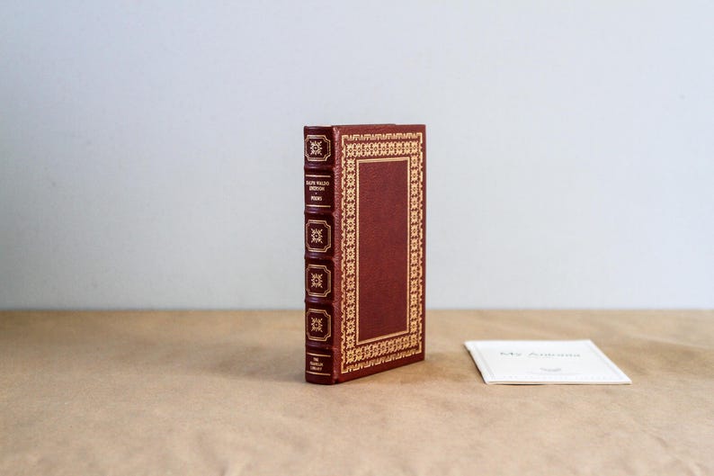 May include: A hardcover book with a burgundy cover and gold decorative accents. The spine features gold lettering and decorative elements. A small white card rests beside the book on a brown surface.
