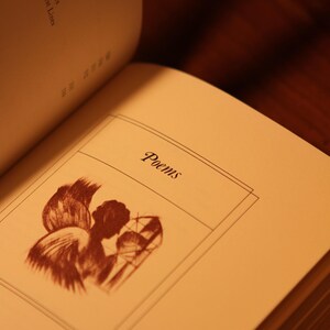 May include: Open book with the title "Poems" on the page. The page features a sepia-toned illustration of an angel with wings and a stained-glass window. The book's index is visible, with titles like "Self-Reliance" and "Index of Titles".