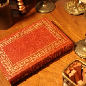 May include: A red leather-bound book with gold decorative trim lies on a wooden surface. The book is surrounded by antique candle holders and a small container of wooden matches, creating a vintage aesthetic.