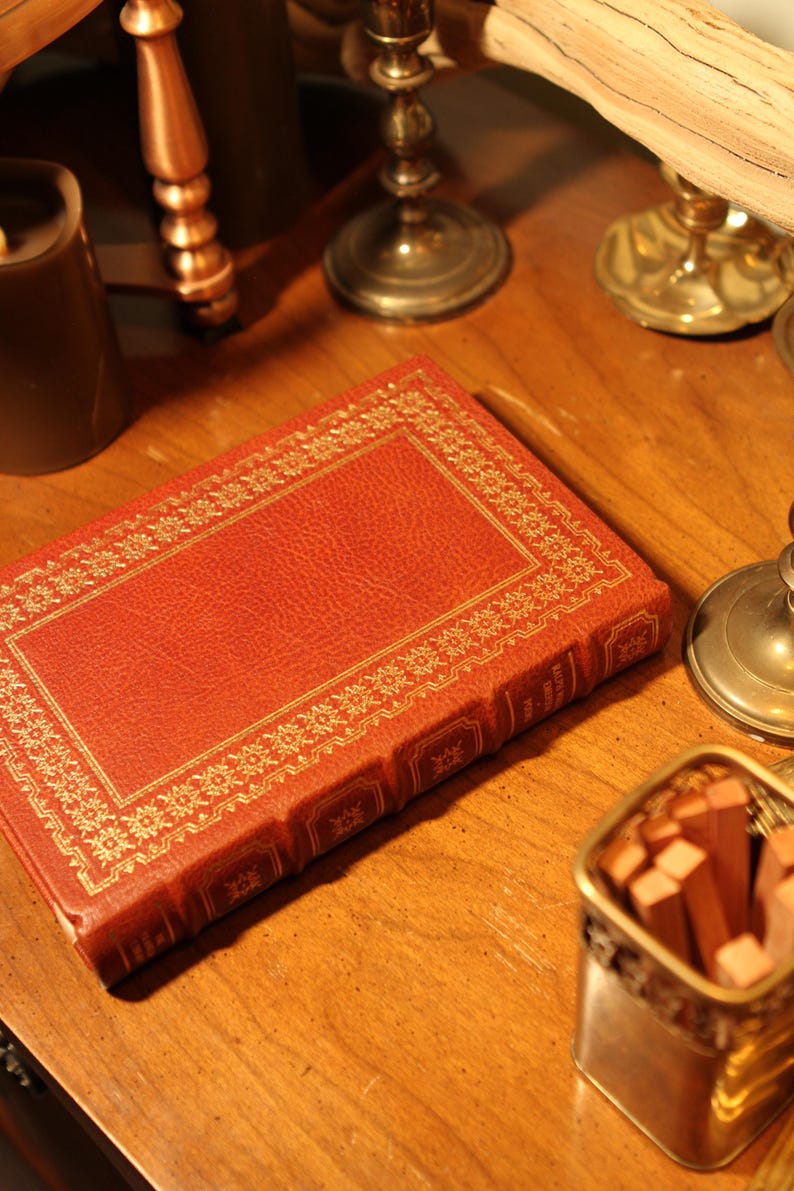 May include: A red leather-bound book with gold decorative trim lies on a wooden surface. The book is surrounded by antique candle holders and a small container of wooden matches, creating a vintage aesthetic.