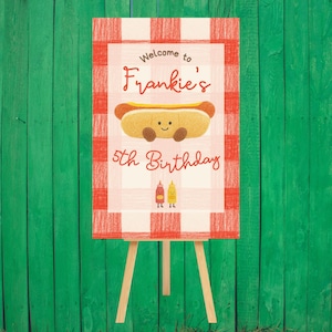 May include: A birthday sign on a wooden easel. The sign has a red and white checkered border and features a cartoon hot dog with a smiling face. Text reads "Welcome to Frankie's 5th Birthday". Two condiment characters are at the bottom.