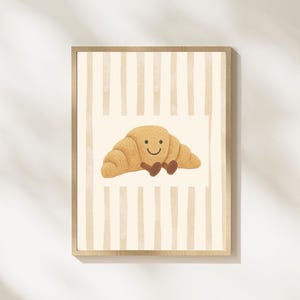 May include: A framed print featuring a cartoon croissant with a smiling face. The croissant is light brown with darker brown details. The background has vertical beige stripes. The frame is made of light-colored wood.
