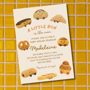 Puede incluir: Invitación a baby shower con el texto "A Little Bun is the Oven" e ilustraciones de productos horneados sonrientes. La invitación es para Madeleine, con fecha, hora y lugar.
