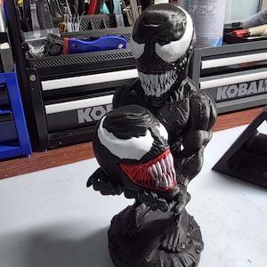 May include: A black and white 3D-printed Venom statue. The statue features a detailed depiction of the character's head and body, with white eyes and teeth, and a red tongue. The statue is on a round base.