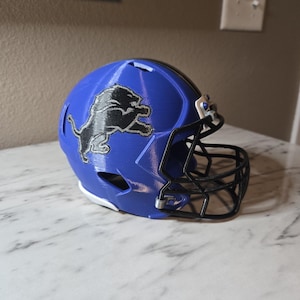 Peut inclure: Casque de football américain bleu avec le logo des Lions de Detroit. Le casque a un masque noir et une bande noire. Le logo est un lion argenté. Le bas du casque est blanc.