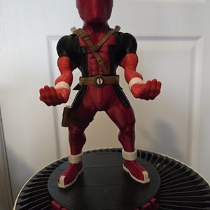 May include: A red and black Deadpool figurine with white eyes and a mask. The figure is standing on a black and red circular base. The figure is wearing a brown belt and a brown strap across the chest.