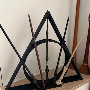 May include: A black triangular wand display stand holding six wands of varying colors and designs. The wands are brown, black, and tan. The stand is on a light-colored wooden shelf.