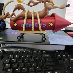 May include: A detailed figurine of Wile E. Coyote riding an ACME rocket. The rocket is red with yellow stars and the text "ACME CO." in blue. The coyote is brown and tan, with a lit fuse and rope detailing.