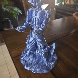May include: A blue figurine of a muscular character in a dynamic pose, with spiky hair and detailed clothing. The figure is standing on a textured base. The figurine is a collectible item.