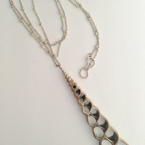 Pode incluir: Um colar de prata com uma corrente delicada e um grande pingente de concha aberta. O pingente é feito de prata e tem um design em espiral.