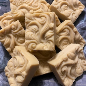 May include: A pile of light beige, square-shaped soaps with a swirling, ornate design carved into each bar. The soaps are stacked on a blue patterned fabric, creating a textured and visually appealing arrangement.