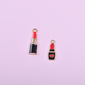Op de afbeelding: Twee lippenstiftbedels. Eén is zwart met een gouden basis en een rode lippenstift, versierd met strass. De andere is zwart met een rode lippenstift en een hartontwerp. Beide hebben gouden lussen om op te hangen.