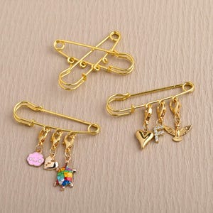 May include: Gold-colored safety pins with attached charms. One pin features a cloud, seashell, and turtle charm. Another has a heart, letter "F", and dove charm. The third pin is a double safety pin.