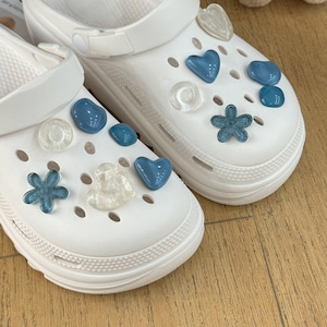 Peut inclure: Sabots blancs décorés de breloques bleues en forme de cœur et de fleur, et de breloques transparentes rondes et en forme de cœur. Les sabots sont en matière lisse et caoutchouteuse.