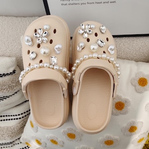 May include: A pair of beige clogs with a pearl and rhinestone embellishment. The clogs have a rounded toe and a thick sole.