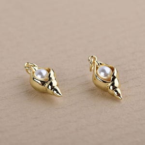 May include: Two gold-colored seashell charms, each with a white pearl nestled inside. The charms have a loop at the top for attaching to jewelry. The seashell design has a textured surface and a pointed tip.