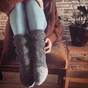 May include: A pair of gray knitted slipper socks with white trim. The socks are worn by a person sitting on a brown bench.