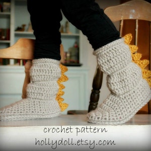 May include: Crocheted dinosaur slippers with a gray body and yellow spikes. The slippers are worn by a person standing on a wooden floor. The text "crochet pattern hollydoll.etsy.com" is printed below the slippers.