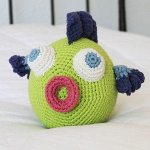 May include: A green crocheted fish toy with blue and white eyes, a pink mouth, and blue fins.