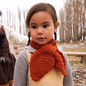 May include: A young girl with brown hair wearing a brown knitted scarf and a light brown dress. She is standing in front of a blurred background of trees.