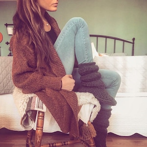 May include: A woman wearing a brown cardigan, blue jeans, and gray knitted leg warmers. She is sitting on a wooden stool with a white fuzzy blanket.