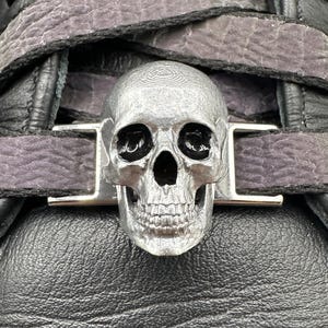 Puede incluir: Primer plano de un zapato de cuero negro con una hebilla de calavera plateada. La hebilla está centrada en la correa del zapato y la calavera tiene detalles. El zapato tiene cuero texturizado y correas grises.