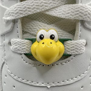 May include: A white trainer with white laces, featuring a yellow cartoon character shoe charm. The charm has black eyes and a green base, adding a playful touch to the footwear.