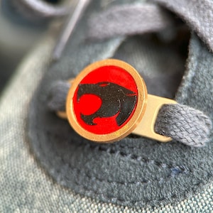 Puede incluir: Un abalorio de cordón de zapato de color dorado con una imagen roja y negra de una cabeza de pantera gruñendo. El abalorio está unido a un cordón de zapato gris.