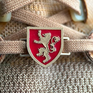 Puede incluir: Un charm de zapato de metal dorado en forma de escudo con fondo rojo y un león dorado. El león está de pie sobre sus patas traseras con la cola enroscada alrededor de su cuerpo.