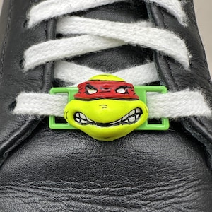 Puede incluir: Primer plano de una zapatilla de cuero negro con cordones blancos. Un dije de zapato de la Tortuga Ninja Mutante Adolescente amarillo y verde está enhebrado a través de los cordones. El dije presenta una máscara roja y una cara sonriente.