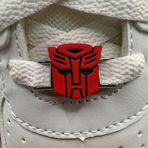 May include: Close-up of a white sneaker with a red and black Transformers logo shoe charm. The charm is rectangular and attached to the laces with a silver metal bar. The laces are white.