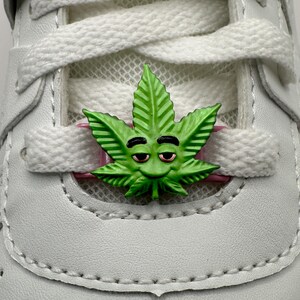 Puede incluir: Un dije de zapato en forma de hoja de cannabis verde con una cara sonriente y detalles en rosa. El dije está colocado en los cordones blancos de una zapatilla blanca. El dije añade un toque único al calzado.