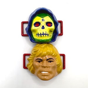 Op de afbeelding: Twee plastic figuren van populaire personages uit de Masters of the Universe franchise. De bovenste figuur is Skeletor, met een blauw en groen schedelhoofd, en de onderste figuur is He-Man, met een geel en bruin hoofd.