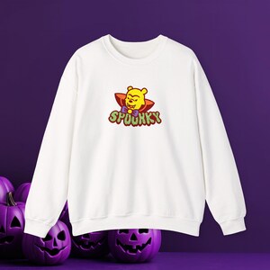 Puede incluir: Una sudadera blanca de manga larga con un gráfico de un oso amarillo vestido de vampiro con pajarita morada y capa roja. Debajo del personaje, la palabra SPOOKY está impresa en una fuente verde, burbujeante y con textura.