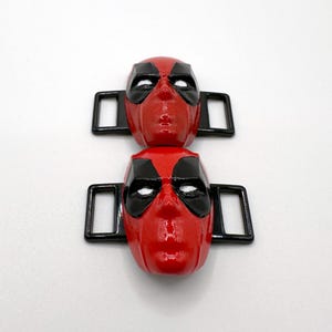以下が含まれることがあります： 赤と黒のデッドプールのマスク型アクセサリーが2つ。マスクには黒い目の覆いがあり、黒い長方形のフレームに取り付けられています。マスクは鮮やかな赤色で、プラスチックまたは樹脂でできている可能性があります。