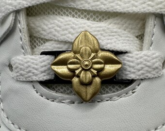 May include: A close-up shot of a white trainer featuring white laces and a gold-coloured floral embellishment. The decorative accent is positioned centrally on the laces, enhancing the shoe's design.
