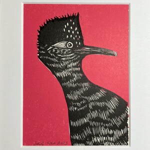 May include: A black and white linocut print of a bird with a long beak and a crest on its head. The bird is printed on a bright pink background. The print is signed "Julie Sola 2023" in the bottom left corner.