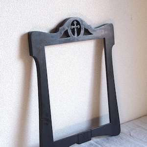 May include: A dark gray wooden frame with a decorative top featuring a carved cross. The frame has a rectangular shape with curved edges and a small, rectangular base. The frame is leaning against a textured wall.