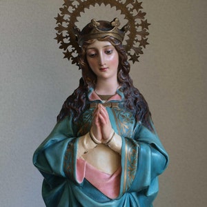K&ouml;nnte beinhalten: Statue einer Frau mit Krone und Heiligenschein, die ein blaues Gewand mit goldenen Akzenten und ein rosa Untergewand tr&auml;gt. Ihre H&auml;nde sind zum Gebet gefaltet. Die Statue hat einen friedlichen Gesichtsausdruck.