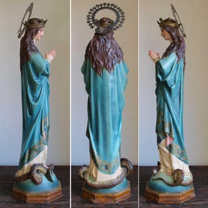 K&ouml;nnte beinhalten: Vintage Statue einer Frau in einem flie&szlig;enden tealfarbenen Gewand, geschm&uuml;ckt mit einer goldenen Krone und Heiligenschein. Die Statue steht auf einem Sockel mit einer sich windenden Schlange. Die H&auml;nde der Figur sind zum Gebet gefaltet.